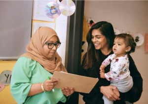 Indoors, a woman in a tan hijab, glasses, and a bright green top holds a clipboard and a green marker, looking down at a paper. Standing next to her, another woman in a dark sweater smiles as she holds a toddler wearing a floral long-sleeved shirt and grey pants. CDs hang from the ceiling above them in what appears to be a classroom or community space.