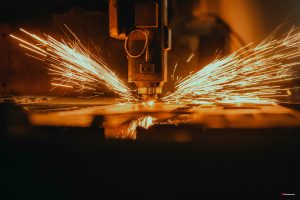 Close-up view of an automated laser cutting machine cutting metal in a dark industrial environment. Bright orange sparks burst outward in both directions as the intense laser head processes the metal sheet.